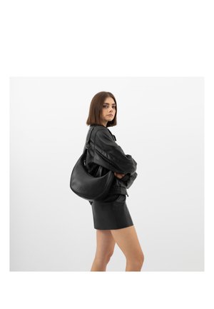 Junge Frau trägt eine schwarze Lederjacke und einen Rock, mit einer großen schwarzen Lederhandtasche über der Schulter, seitlich stehend.