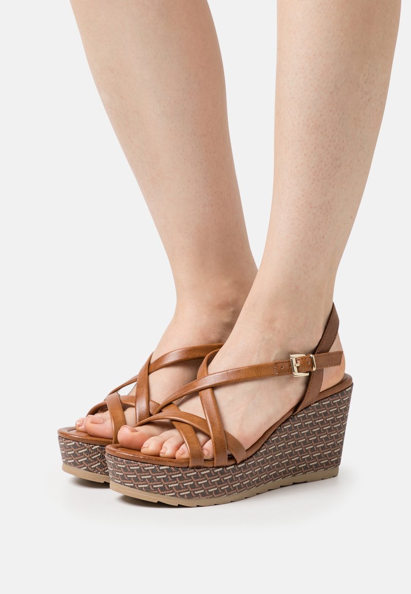 Pieds portant des sandales compensées en cuir marron avec des brides croisées et une boucle, dotées d'une semelle plateforme à motifs.