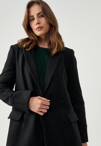Černý blazer z vlněné směsi s klopovým límcem, zapínáním na jeden knoflík a bočními kapsami. Nesený přes žebrovaný zelený svetr.