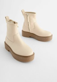 Beige Leder-Ankle-Boots mit einer klobigen, strukturierten Sohle, seitlichem Reißverschluss und Zuglasche, die sich durch klare Linien und ein abgerundetes Design auszeichnen.