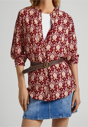 Femme portant un chemisier boutonné à motifs bordeaux avec une ceinture marron sur un haut blanc et une mini-jupe en jean bleu.