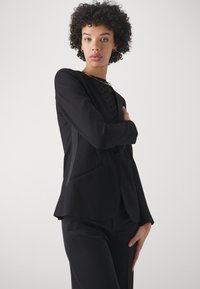 Pielāgota melna žakete ar rakstainu blūzi zem tās, kurai ir šauras atloks un divas sānu kabatas. Elegants un strukturēts dizains.