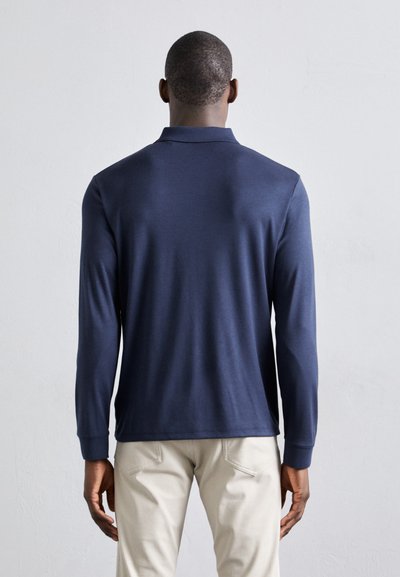 Ναυτικό μακρυμάνικο πόλο μπλουζάκι με κλασικό γιακά, μαλακή υφή και εφαρμοστή σιλουέτα. Συνδυάζεται με ανοιχτόχρωμο παντελόνι για αντίθεση.