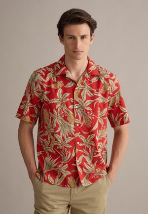 Joven con una camisa roja de manga corta con botones y patrones florales beige, y pantalones beige, de pie frente a un fondo liso.