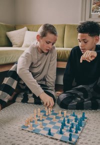 Skakspil i gang på et ternet bræt. Træbrikker i lyse og mørkeblå nuancer, med spillere iført ternede bukser, fokuseret på brættet.