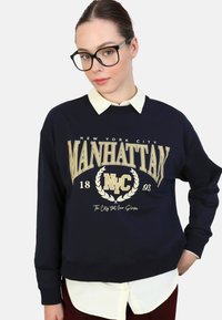 Felpa navy con la scritta "MANHATTAN NYC" stampata in oro, una camicia bianca con colletto sotto e una vestibilità comoda. Tessuto morbido e polsini a coste.