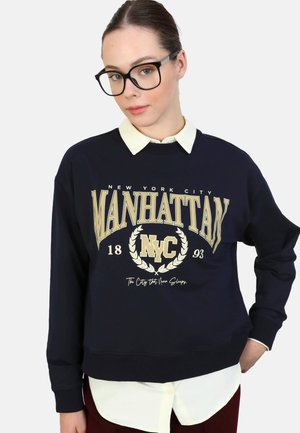 Sudadera de marina con la impresión en dorado "MANHATTAN NYC", con una camisa de cuello blanco debajo y un corte relajado. Textura de tela suave y puños acanalados.