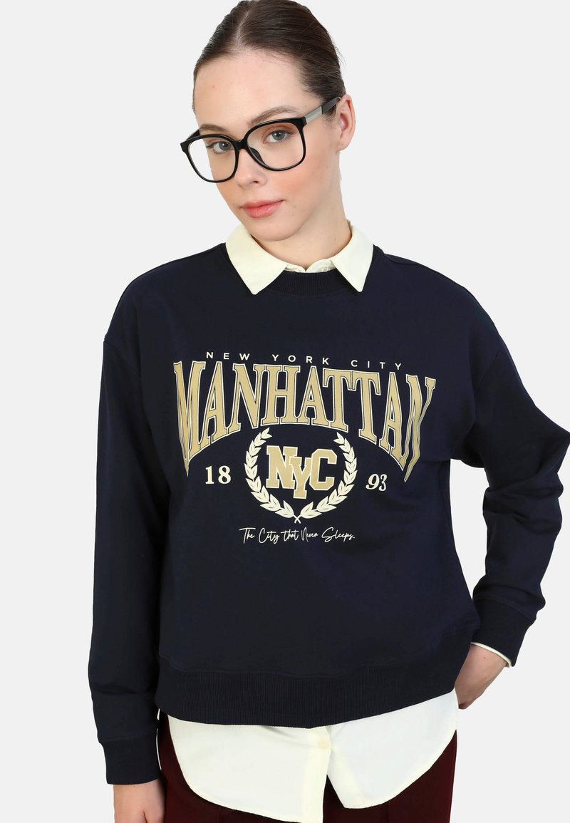 Felpa navy con la scritta "MANHATTAN NYC" stampata in oro, una camicia bianca con colletto sotto e una vestibilità comoda. Tessuto morbido e polsini a coste.