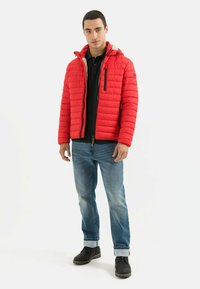 Rote, wattierte Jacke mit horizontalem Steppdesign, abnehmbarer Kapuze, Frontreißverschluss und schwarzen Akzenten. Getragen mit blauen Jeans und schwarzen Stiefeln.
