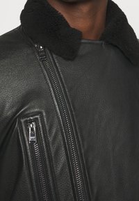 Svart läderjacka med shearlingkrage, med dragkedjor framtill och på sidan, samt en texturerad yta på materialet.