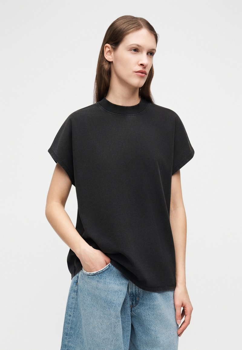 Femme vêtue d'un haut noir ample sans manches et d'un jean en denim bleu clair, debout avec une main dans la poche devant un fond uni.