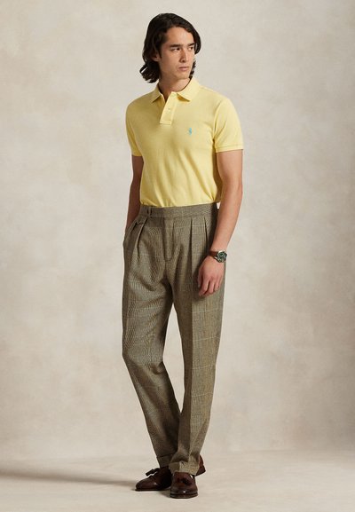 Vyras dėvi geltoną trumpomis rankovėmis polo marškinėlį, rudus languotus klostuotus kelnes, rudus kutuoto stiliaus mokasinus ir laikrodį ant riešo, stovintis rankomis kišenėse.