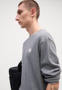 Γκρίζο πουκάμισο με μακριά μανίκια σε πλέγμα waffle με μικρή ετικέτα λογότυπου. Μαύρη υφασμάτινη τσάντα κρατημένη στο άλλο χέρι, παρουσιασμένη σε ουδέτερο φόντο.