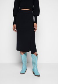 Jupe noire, mi-longue jusqu'aux genoux, moulante avec un design portefeuille. Associée à des bottes de cow-boy bleu clair avec des coutures détaillées et un bout pointu.