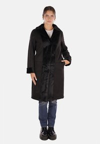 Cappotto nero con una texture simile alla pelliccia nella parte anteriore, maniche lunghe e due tasche laterali. Il modello indossa jeans di denim blu e scarpe nere.