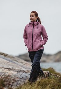 Light pink waterproof jacket with a hood, featuring black zipper and drawstrings; paired with black pants in a rugged outdoor setting.