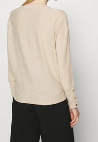 Beiges Strickoberteil mit langen Ärmeln, strukturiertem Muster, abgerundetem Saum und drei Knopfakzenten an den Ärmelbündchen.