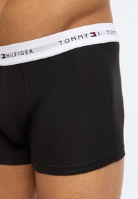 Άνδρας με κορμί που φοράει μαύρα boxer Tommy Hilfiger με λευκή ζώνη μέσης και λογότυπο.