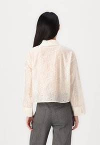 Cropped, sheer white blouse with floral embroidery and open stitching details. Features a collar and long sleeves with cuff accents.