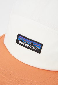 Baseball cap featuring a white body and an orange visor. Embroidered black Patagonia logo with colorful mountain design.  Cotton material.