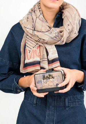Beige Schal mit abstraktem Muster, der um eine Jeansjacke gewickelt ist. Eine gemusterte Geldbörse mit einem schwarzen Grund, die in den Händen gehalten wird und ein verspieltes Design aufweist.