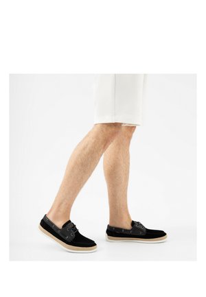 Benedenbenen met witte korte broek en zwarte instapschoenen met beige zolen, lopend op een effen witte achtergrond.