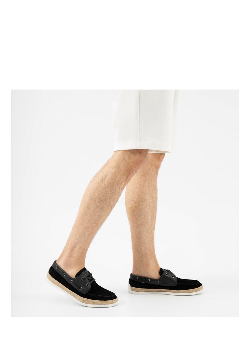 Nohy oblečené v bílých šortkách a černých slip-on casual lodních botách s béžovými podrážkami, chodící na jednobílém pozadí.