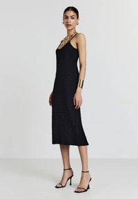 Schwarzes Midi-Kleid aus strukturiertem Stoff, mit dünnen Trägern und einem runden Ausschnitt. Kombiniert mit schwarzen, schnürenden High Heels und einer goldenen Statement-Halskette.