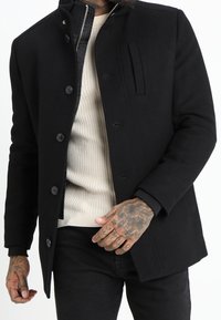 Man med svart knappad kappa över en beige ribbad tröja, som visar tatuerade händer och en delvis dragkedjad inre jacka.