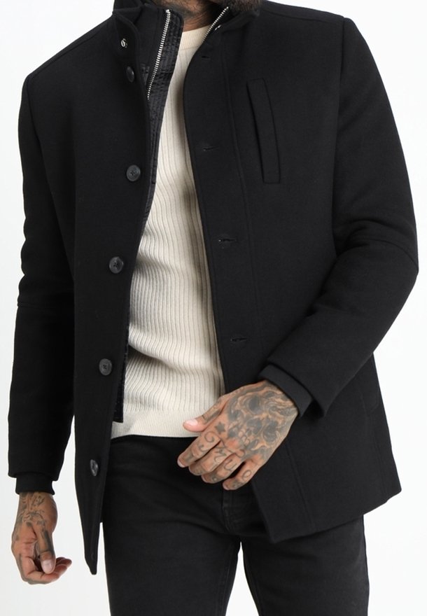 Man med svart knappad kappa över en beige ribbad tröja, som visar tatuerade händer och en delvis dragkedjad inre jacka.