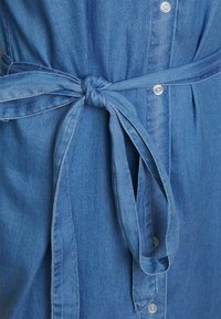 Vestido de ganga em azul claro, com um cinto amarrado na cintura, fecho frontal com botões e tecido suave e texturizado. Design casual com mangas dobradas.