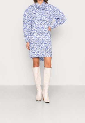 Kvinde iført en blå og hvid blomsterprintet langærmet kjole med en stor sløjfe ved halsen og knæhøje cremefarvede støvler.