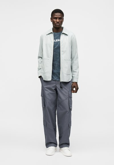 Άνδρας φορώντας ανοιχτό γκρι μπουφάν πάνω από σκούρο μπλε μπλουζάκι, γκρι παντελόνια cargo και λευκά αθλητικά παπούτσια, στέκεται μπροστά σε απλό λευκό φόντο.