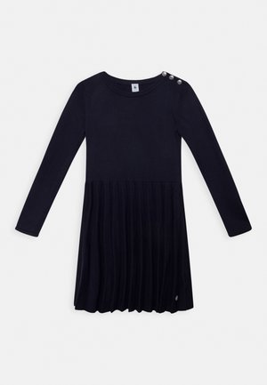 Czarna sukienka z długim rękawem i plisowaną spódnicą, okrągłym dekoltem i trzema metalowymi guzikami na lewym ramieniu.