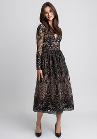 Czarna koronkowa sukienka z kwiatowymi wzorami, długimi rękawami, głębokim dekoltem w kształcie V oraz przewężoną talią. Uzupełniona przezroczystą podszewką i lekko opadającą spódnicą.