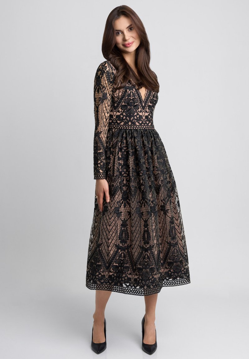 Czarna koronkowa sukienka z kwiatowymi wzorami, długimi rękawami, głębokim dekoltem w kształcie V oraz przewężoną talią. Uzupełniona przezroczystą podszewką i lekko opadającą spódnicą.