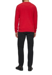 Sarkans adīts džemperis ar garām piedurknēm, apaļu kakla izgriezumu un ribotām aprocēm, kombinēts ar melniem džinsiem un melnām kurpēm, skatīts no aizmugures.