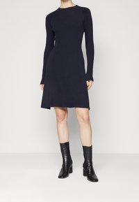 Sukienka dzianinowa z długim rękawem w ciemnym granacie, z dopasowaną górą i rozkloszowanym dołem, zestawiona z czarnymi skórzanymi botkami za kostkę. Gładka faktura i prosty design.