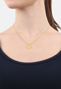 Guldig halsband med ett hjärtformat hänge och graverade detaljer, som hänger på ett delikat kedja, bärs mot en mörkblå topp.