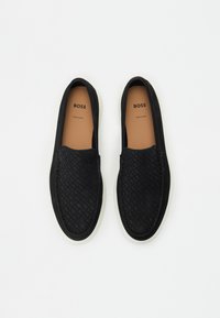Melni slip-on apavi ar austiem rakstiem purngalā, gluda zamšāda, balta gumijas zole un zīmola brūns iekšzole.