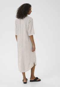 Abito a righe di lunghezza media in tessuto leggero con linee verticali, maniche corte, fessure laterali e vestibilità rilassata. Indossato con sandali neri tipo slide.