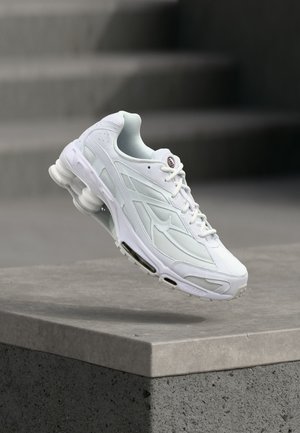 White athletic sneaker with mesh and leather details floating above a concrete ledge with blurred gray background.