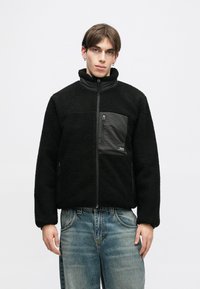 Giacca in pile nera con colletto alto e tasca sul petto con zip. Presenta un tessuto morbido e testurizzato e un pannello liscio a contrasto nella parte anteriore.