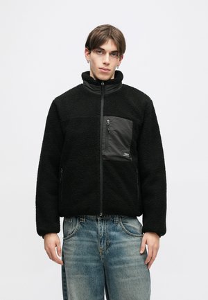 Čierna fleecová bunda s vysokým golierom a zipsovou náprsní kapsou. Je vybavená textúrovanou, mäkkou tkaninou a kontrastným hladkým panelom na prednej strane.