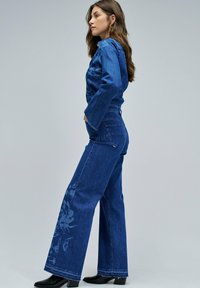 Mono de mezclilla en azul oscuro, con piernas anchas y patrones florales en la pierna izquierda. Cuello clásico y cierre con botones en la parte delantera, con mangas largas.