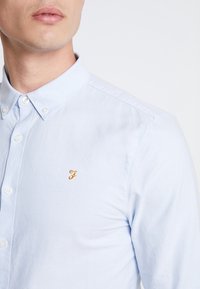 Camisa de botones azul claro, de material de algodón, con cuello. Presenta un pequeño logo bordado en oro con la letra "F" en el lado izquierdo. Diseño clásico.