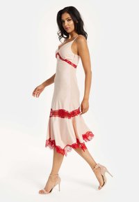 Blush-colored dress with red lace accents, fitted bodice, and flared hem. Adjustable straps. Worn with nude high-heeled sandals.