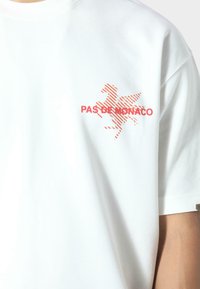 Camiseta de algodón blanca con un gráfico rojo de un caballo alado y el texto "PAS DE MONACO" en letras gruesas. Diseño sencillo de mangas cortas.