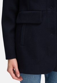 Kvinde iført mørkeblå frakke med klaplomme og sidelomme, vist fra midt på låret til håndleddet, parret med blå jeans.