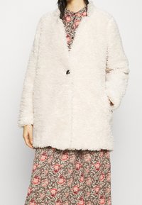 Fluffy, cremefarvet frakke med løs pasform, enkelt knaplukning og to sidelommer. Bæres over en kjole med blomstermønster.